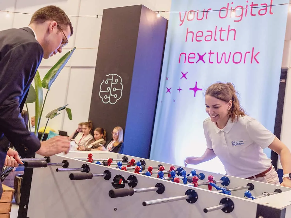 Zwei spielende Personen am Kickertisch in Action. Im Hintergrund ein Plakat mit der Aufschrift: Your digital health nextwork.