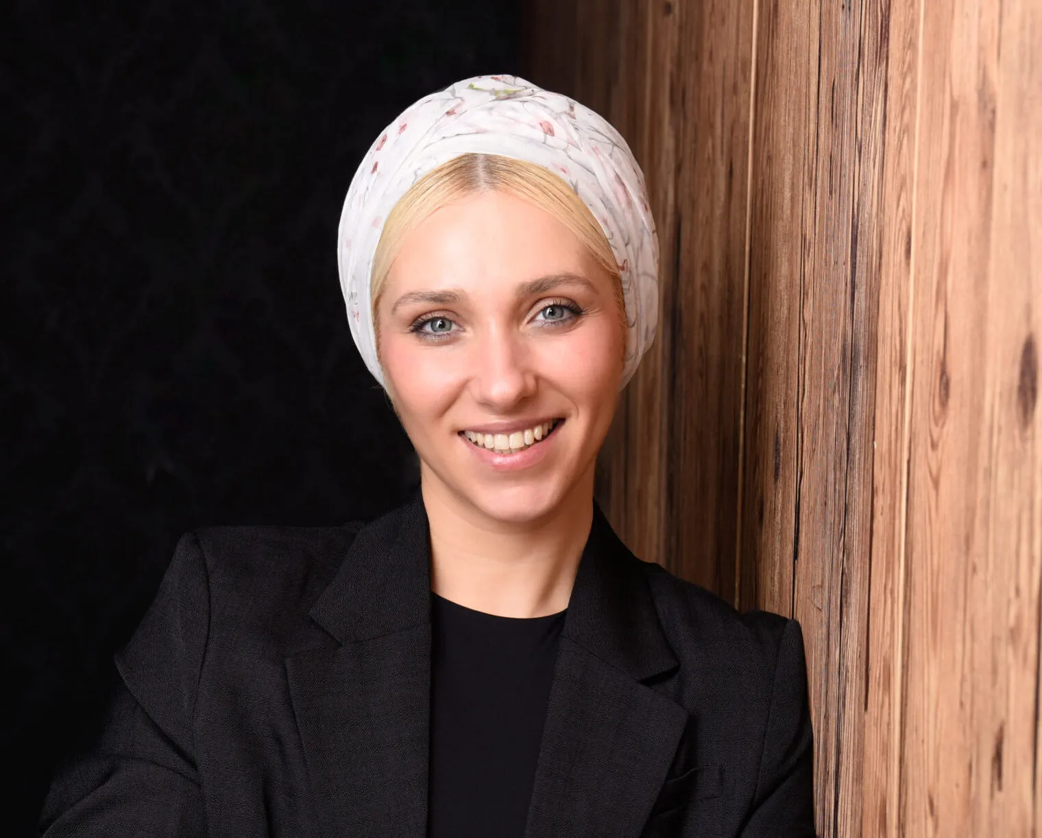 Profilbild von Hanna Hryhorova mit verschränkten Armen lehnt sie sich an eine Holzwand. Trägt ein schwarzen Blazer, lächelt in die Kamera und hat ein Tuch auf Ihren Kopf.