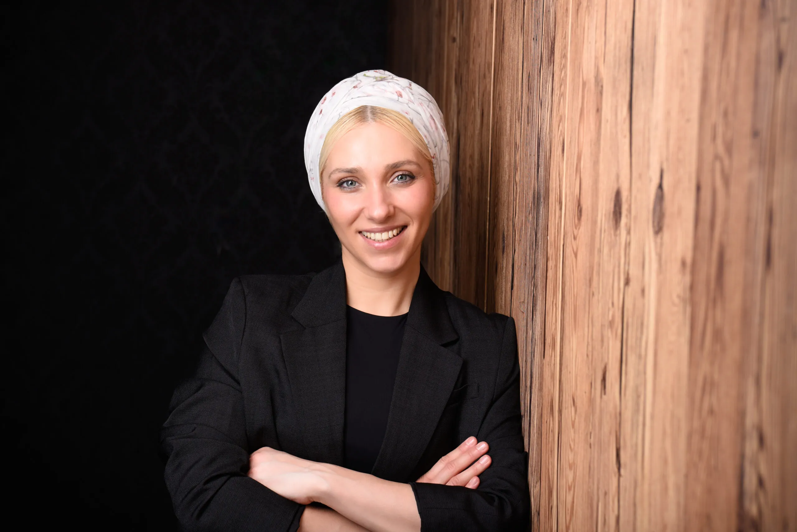 Profilbild von Hanna Hryhorova mit verschränkten Armen lehnt sie sich an eine Holzwand. Trägt ein schwarzen Blazer, lächelt in die Kamera und hat ein Tuch auf Ihren Kopf.