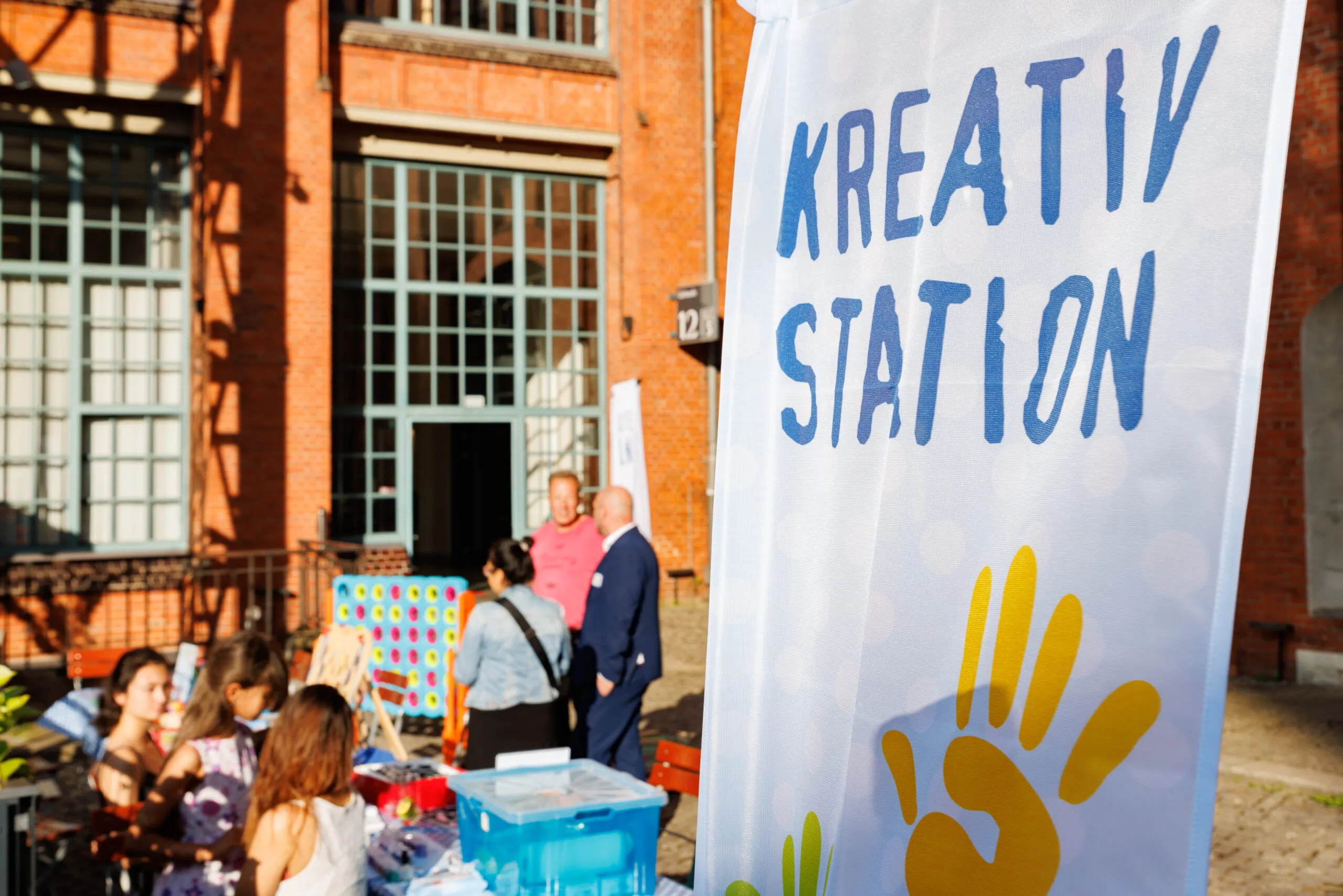 Bild mit einem Plakat mit dem Titel Kreation Station. Im Hintergrund sind stehende und sitzende Menschen zu sehen.