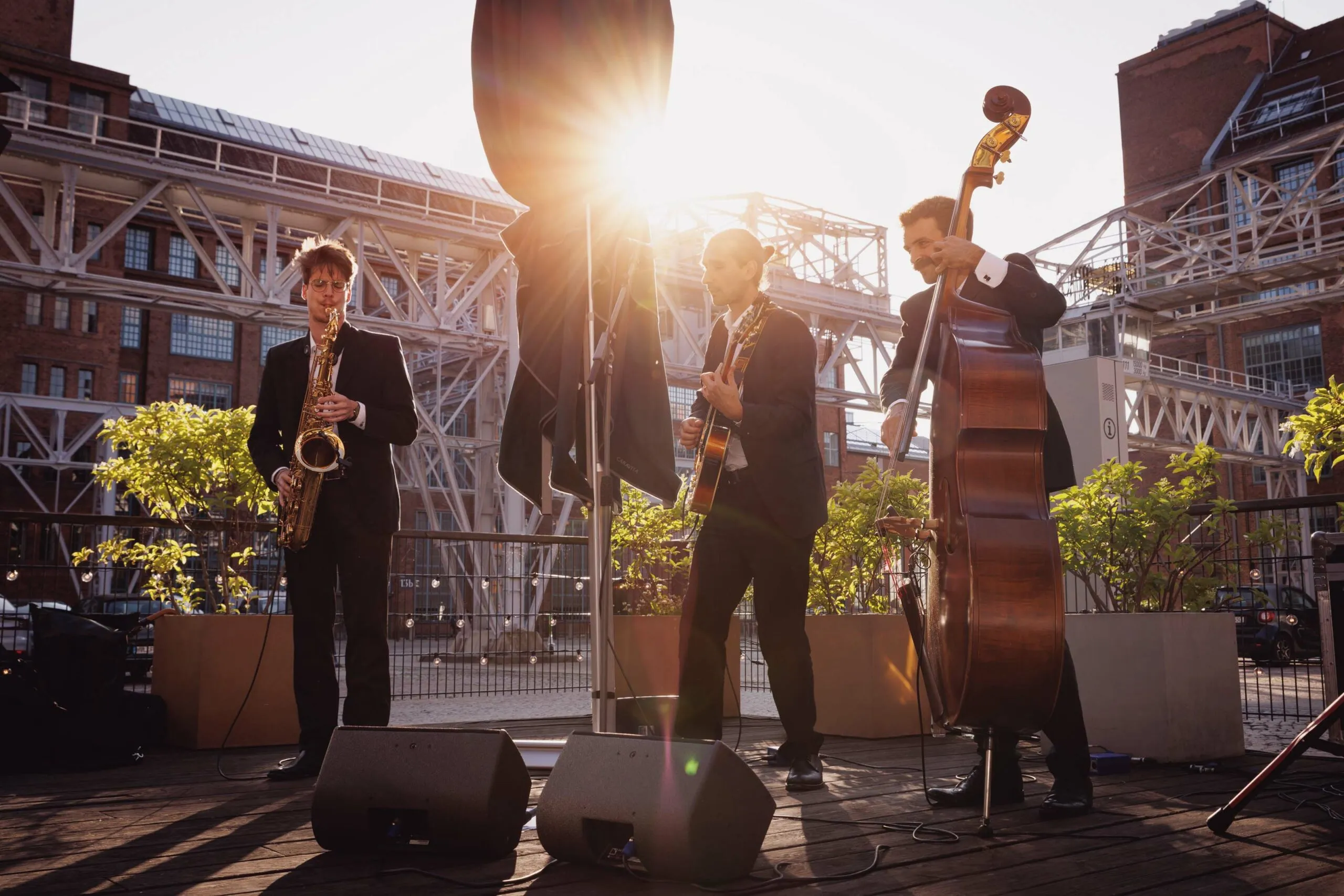 Eine Band aus drei Herren spielen auf verschiedenen Instrumenten in der Abendsonne eines Industriehofes. Einer spielt auf einem Saxophon, einer auf einer Gitarre und der dritte auf einem Kontrabass.