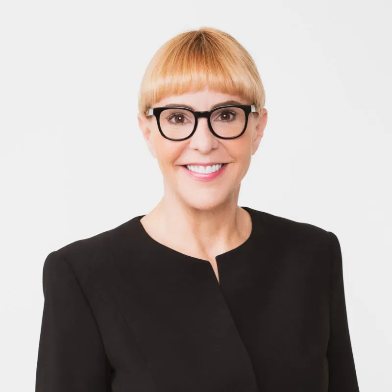 Frau mit schwarzer Brille, lächelt in die Kamer, hat Blonde haare, einen Zwock hinten, den man nicht sieht, einen kurzen Geraden Ponny und einen schwarzen Balzer