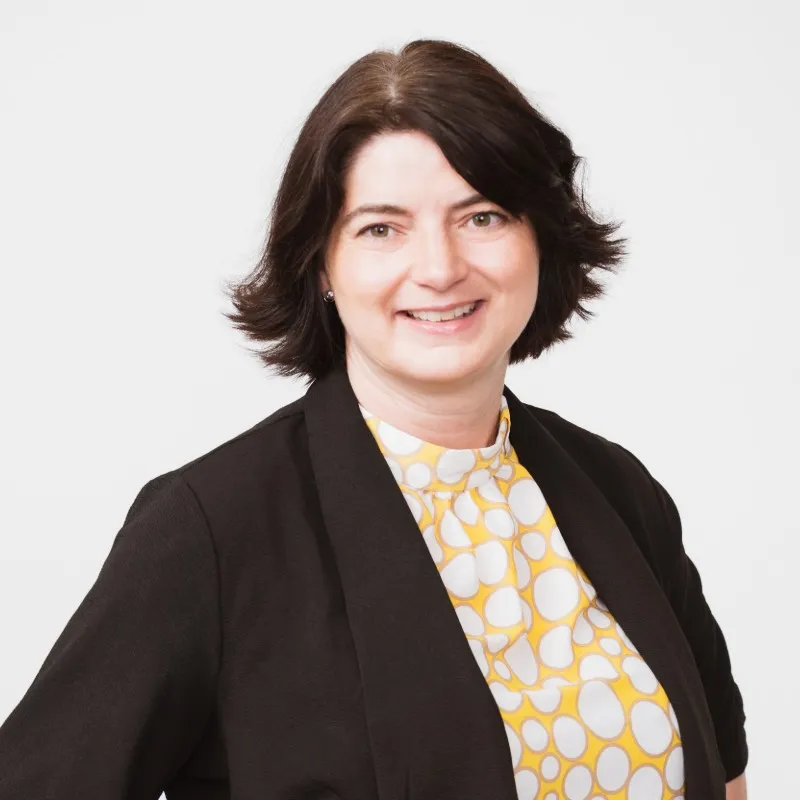 Frau mit gelb-weißen Muster als T Shirt und schwarzem Blazer und kurz dunkelbraunen haaren und lächelt in die Kamera. Ca. 50 Jahre alt.