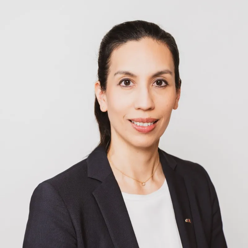 Frau mit Zopf und schwarzen Harren lchelt leicht in die Kamera. hat ein weißes Tshirt und ein schwarzen Blazer an