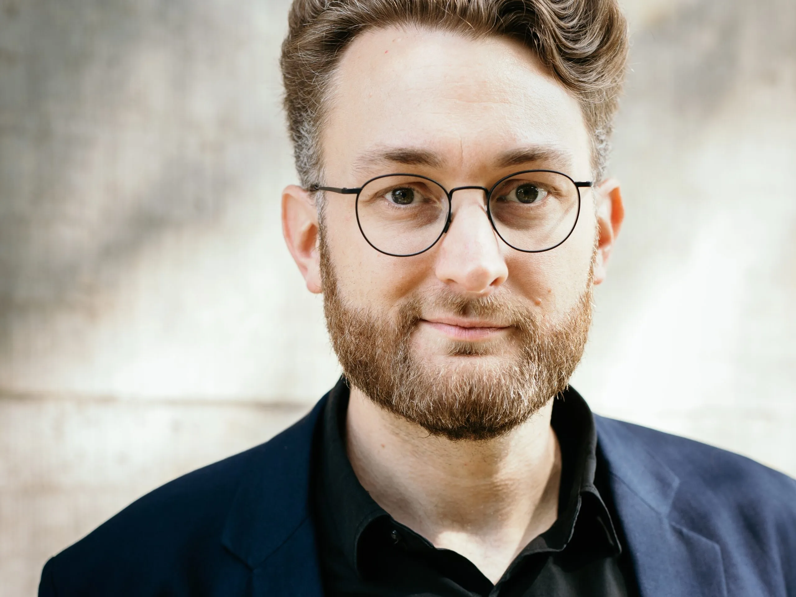 Mann mit Brille um Bart guckt in die Kamera. Trägt ein dunkelblaues Hemd.